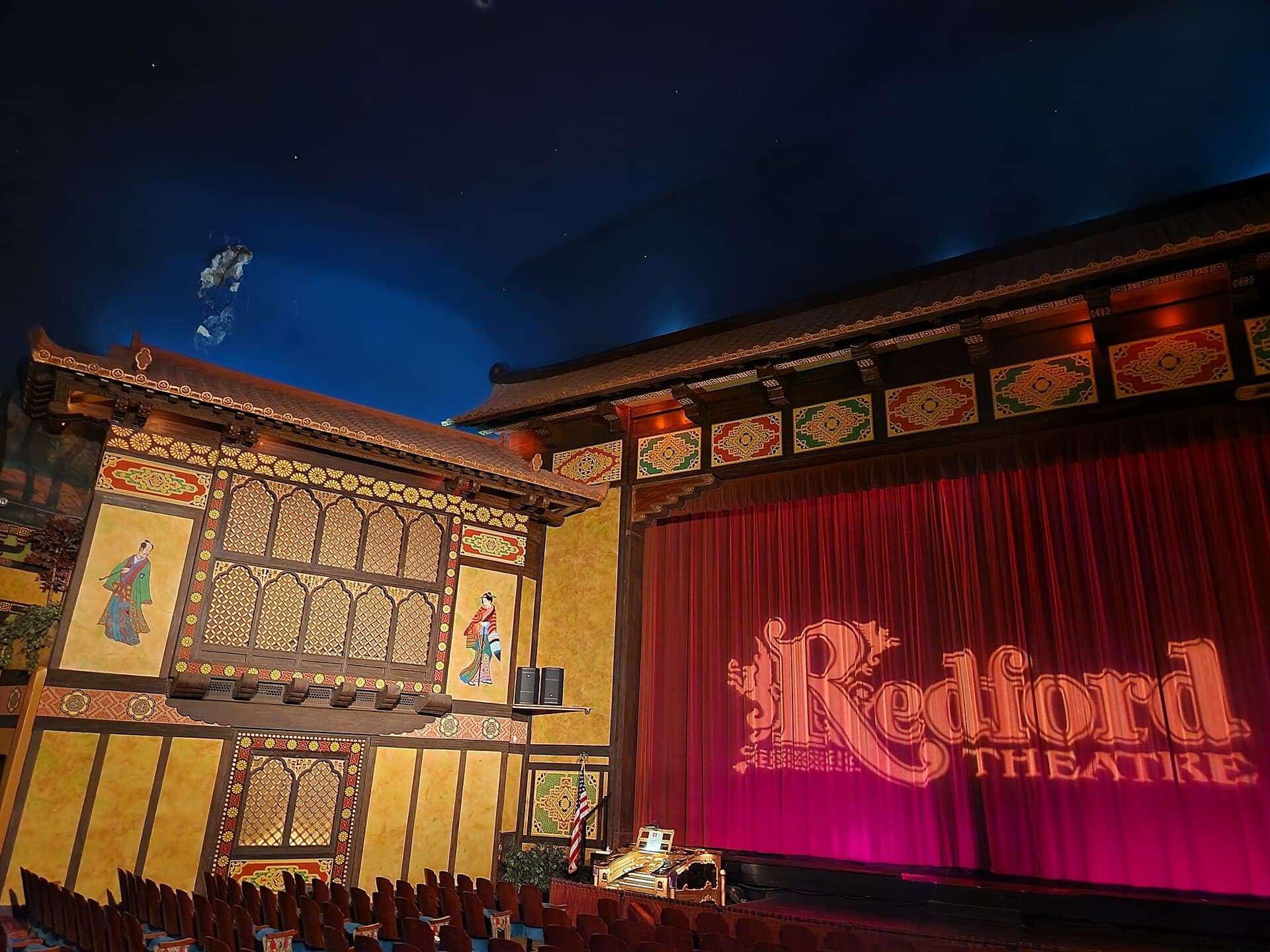 A theatre auditorium with a red curtain and painted proscenium